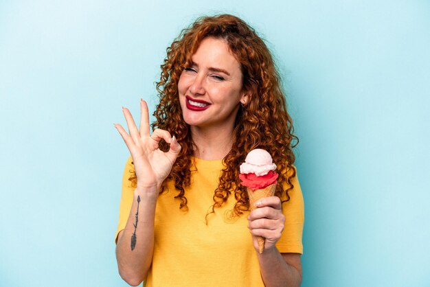 Young ginger woman holding an ice cream isolated on blue background cheerful and confident showing ok gesture