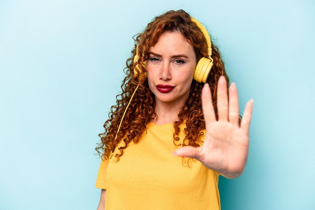 Young ginger woman listening to music isolated on blue background standing with outstretched hand showing stop sign preventing you