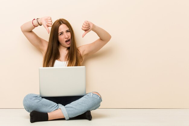 Young ginger woman sitting on her house floor showing thumb down and expressing dislike.