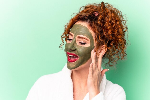 Young ginger woman wearing beauty facial mask isolated on green background