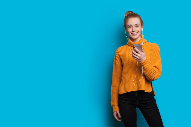 Young ginger woman with freckles is listening to music on a blue wall with free space using headphones