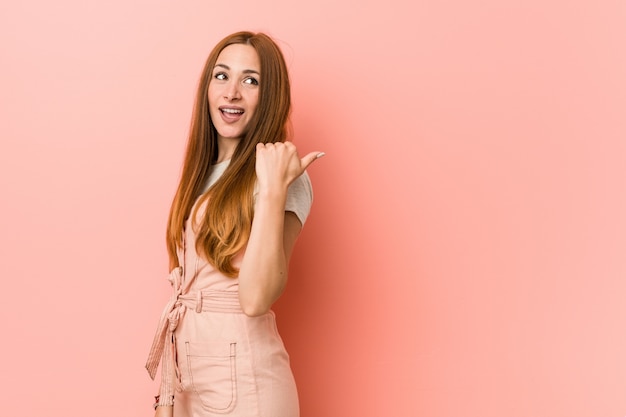 Young ginger woman with freckles points with thumb finger away, laughing and carefree.