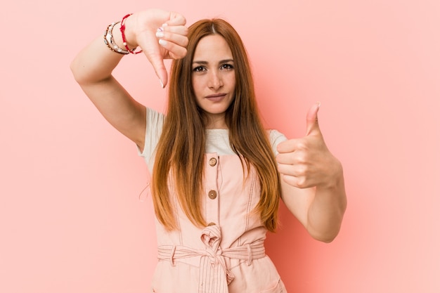 Young ginger woman with freckles showing thumbs up and thumbs down, difficult choose concept