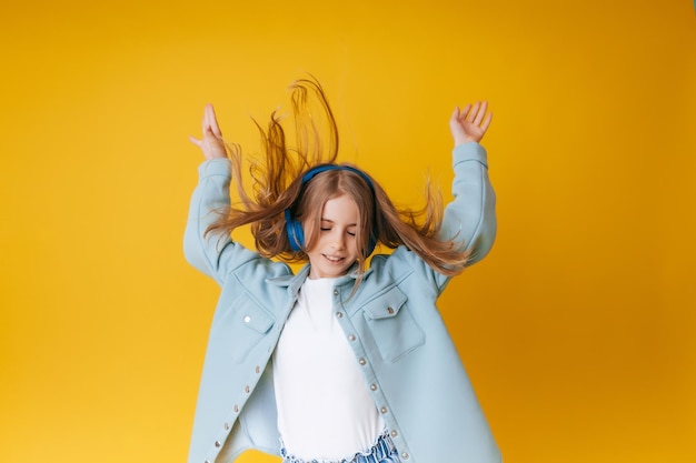 A young girl 1113 years old in headphones listens to music and dances in the studio on a yellow background