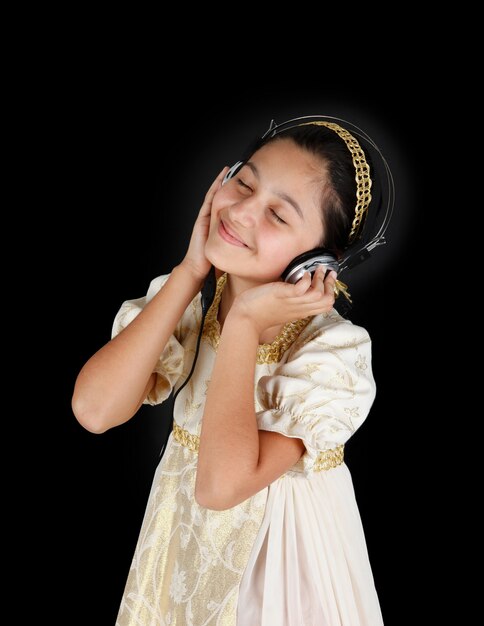 Young girl in ancient dress while listening music with headphones on black wall.