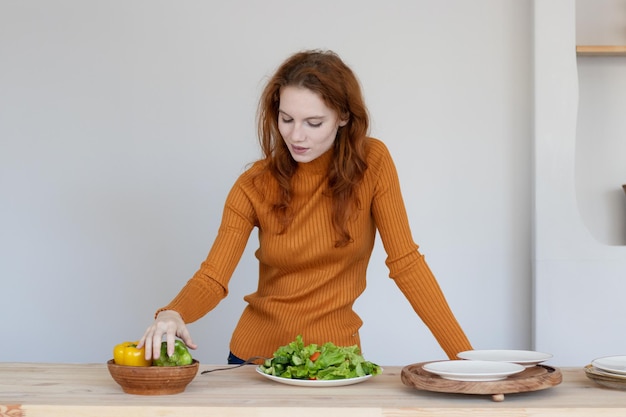 エプロンの少女は、キッチンで野菜とハーブのヘルシーなサラダを作ります