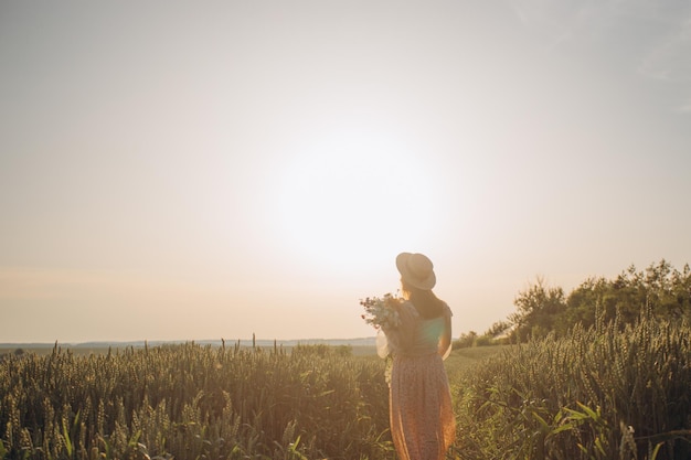 Young girl in a beautiful long dress and hat walks in a field with wheat in his hand