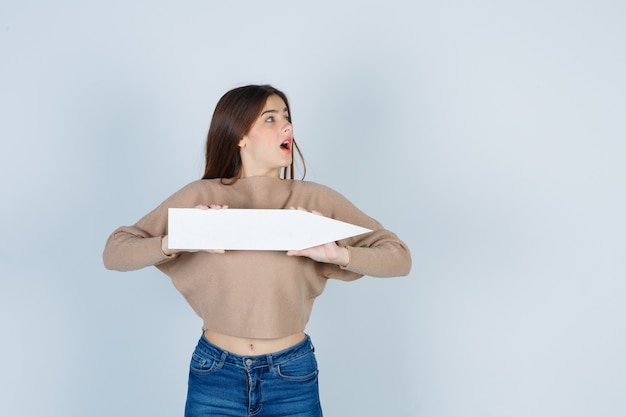 Young girl in beige knitwear, jeans holding paper stick, looking at right side and looking surprised , front view.