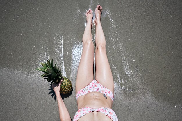 Photo a young girl in a bikini lying on the sand on the beach with a pineapple vacation concept