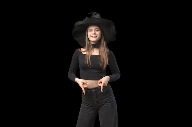 Young girl in black cloth and hat pointing fingers down Teenager in the image of witch on black background Copy space