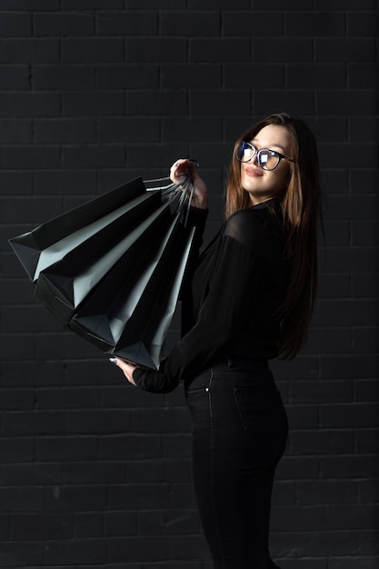 Young girl in black clothes holds shopping bags against black wall Vertical frame