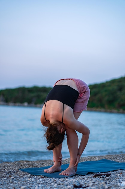 A young girl in a black tank top and pink shorts doing yoga on the sea beach Yoga at sunset overlooking the beautiful Adriatic Sea and green shores Photo promotes a healthy lifestyle sports yoga