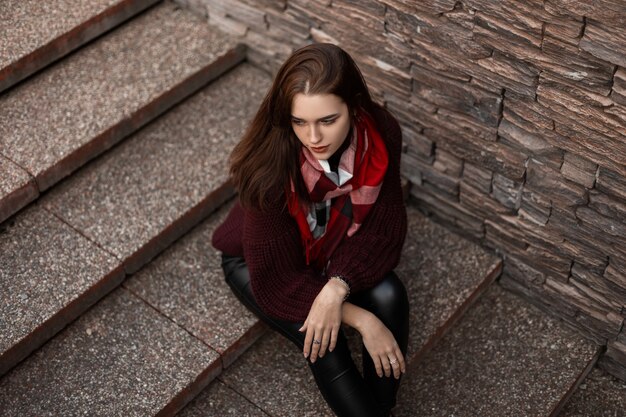 young girl in boots and knitted sweater is sitting in the city on a stone staircase