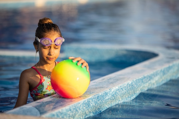 明るいヒョウの水着を着た少女は、暖かい晴れた夏の夜に澄んだ透明な水と深い青色のプールで明るい色の膨脹可能なボールと一緒に泳ぐ