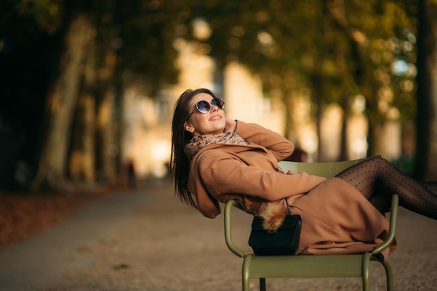 A young girl in a brown coat sits in the middle of the park on a khaki stool girl relax and have good day