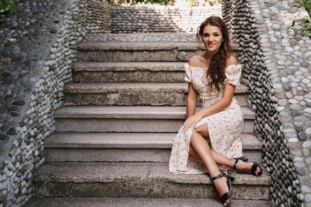 A young girl a brunette with long hair in a vintage dress is sitting on the steps of a gray stone staircase