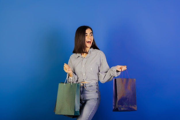 Young girl in casual clothes, blue shirt and jeans holding shopping bags on blue surface.