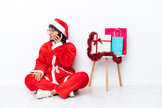 Giovane ragazza che celebra il natale seduta sul pavimento isolato su sfondo bianco mantenendo una conversazione con il telefono cellulare con qualcuno
