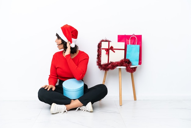 Молодая девушка празднует Рождество, сидя на полу, изолированном на белом bakcground, слушая что-то, положив руку на ухо