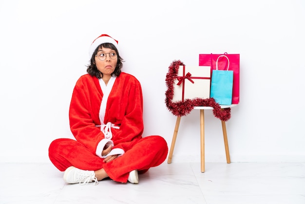 Young girl celebrating Christmas sitting on the floor isolated on white bakcground making doubts gesture while lifting the shoulders