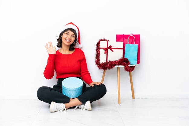 Ragazza che celebra il natale seduta sul pavimento isolato su sfondo bianco che saluta con la mano con espressione felice