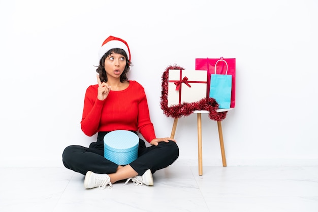 Молодая девушка празднует Рождество, сидя на полу, изолированном на белом bakcground, думая об идее, указывая пальцем вверх