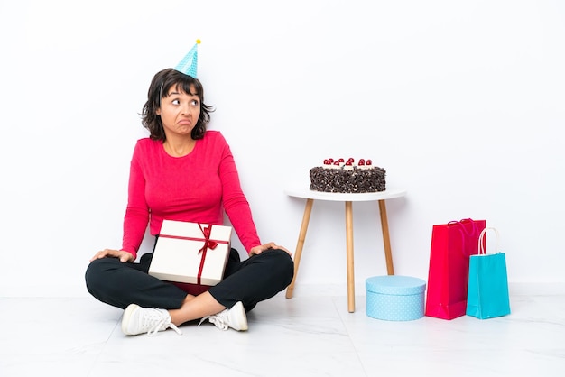 Young girl celebrating his birthday sitting on the floor isolated on white background making doubts gesture while lifting the shoulders