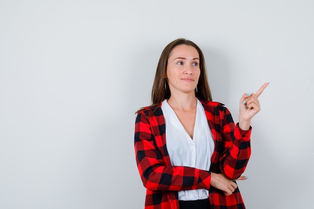 Young girl in checkered shirt, blouse pointing up with finger and looking wistful , front view.