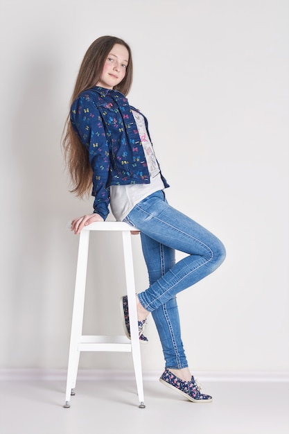 Young girl child sitting on a chair. Smile joy emotions on her face. Girl in jeans posing on light background