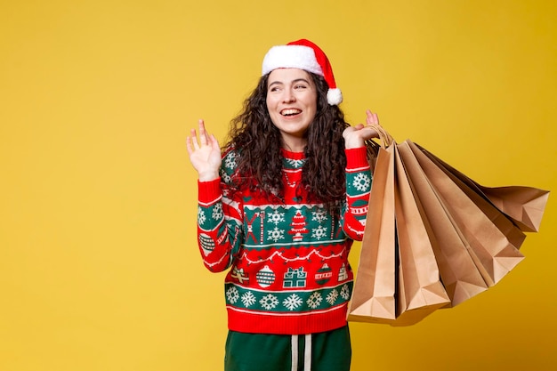 Young girl in christmas clothes and santa hat holds shopping bags and smiles on yellow background