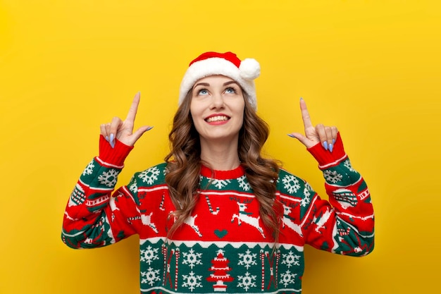 Photo young girl in christmas sweater and santa claus hat smiles and points up at copy space