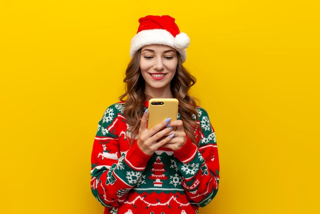 молодая девушка в рождественском свитере и шапке санта-клауса пользуется смартфоном