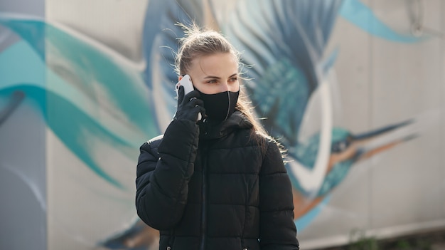 Young girl in city street wearing black sterile medical face mask. Woman using the phone to search for news about nCov 2019. Quarantine COVID-19 pandemic coronavirus epidemic and health care concept