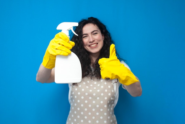 young girl cleaner in uniform holds spray and shows a like on blue background