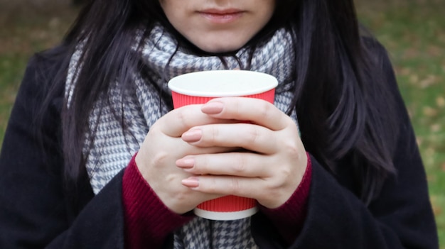 A young girl in a coat holds morning coffee with her while walking in the park. Hand holding paper cup of coffee in green park. Takeaway cappuccino. Close-up.