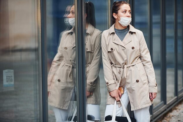 Young girl in coat and white protective mask standing outdoors near building