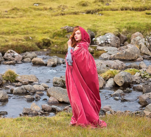 Young girl cosplayer in a red cloak stays near small river in autumn Streymoy Faroe Islands