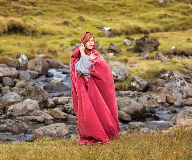 Una giovane cosplayer con un mantello rosso rimane vicino al piccolo fiume streymoy isole faroe