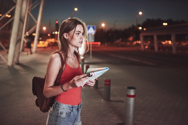 Young girl costs at night near the terminal of the airport or station and reading city map and looking for hotel. Cute tourist with backpacks determine the concept of travel.