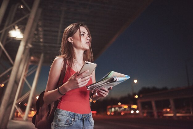 Молодая девушка стоит ночью возле терминала аэропорта или вокзала, читает карту города и ищет отель. Симпатичный турист с рюкзаками определяет концепцию путешествия.