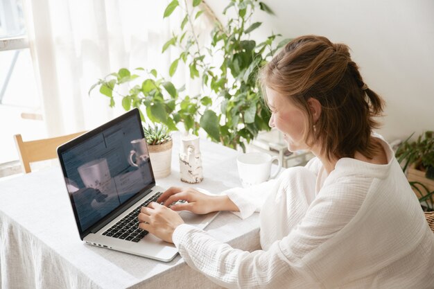 Young girl in cozy white muslin pajamas works on a laptop at home distant work