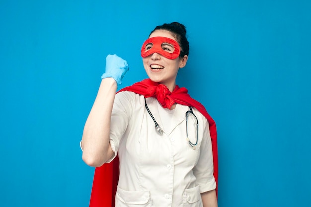 Photo young girl doctor in uniform in superman costume rejoice in victory on blue background