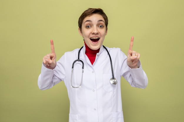 Young girl doctor in white coat with stethoscope around neck happy and surprised showing index fingers standing on green