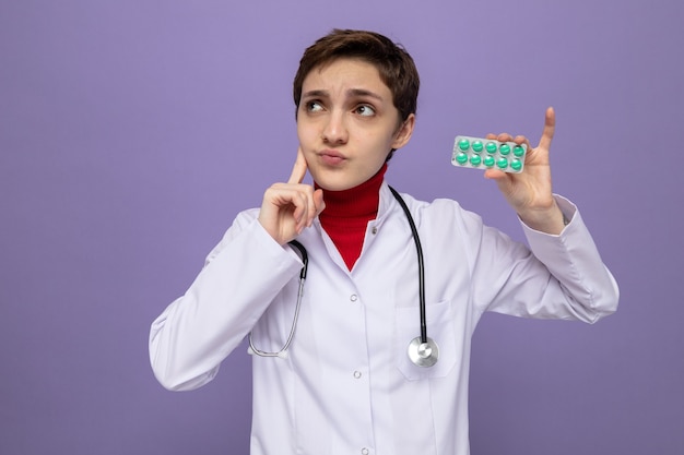 Young girl doctor in white coat with stethoscope around neck holding blister with pills looking up puzzled