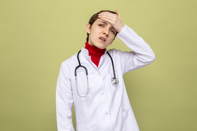 Medico della ragazza in camice bianco con lo stetoscopio intorno al collo che sembra confuso e molto ansioso con la mano sulla sua fronte per errore che sta sopra la parete verde