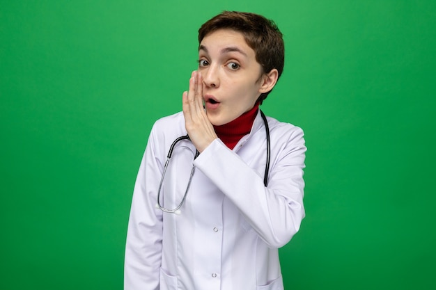 Young girl doctor in white coat with stethoscope around neck telling secret holding hand on mouth standing on green