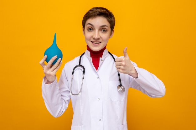 Giovane dottoressa in camice bianco con stetoscopio che tiene in mano una pera medica che sorride mostrando i pollici in piedi sull'arancia Foto Premium