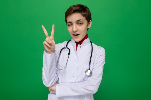 Young girl doctor in white coat with stethoscope smiling confident winking showing v-sign standing on green