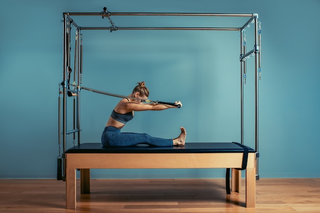 Young girl doing pilates exercises with a reformer bed. Beautiful slim fitness trainer on reformer gray background, low key, art light. Fitness concept