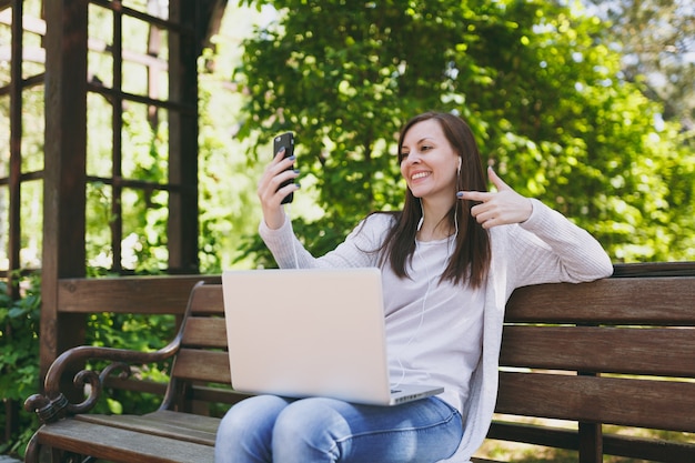 Young girl doing selfie on mobile phone or video call. Woman sitting on bench working on modern laptop pc computer in city park in street outdoors on nature. Mobile Office. Freelance business concept.